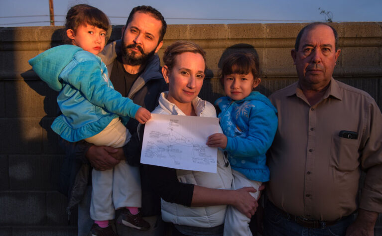 Twin girls with special health care needs are held by their parents while standing next to their grandfather. They are outside, next to a fence, their faces reflecting the sunset. The girls are holding a copy of their care map.