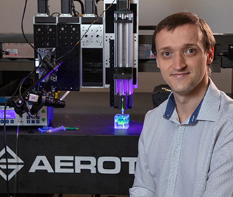 Scientist sitting by 3D printing system, smiling at the camera