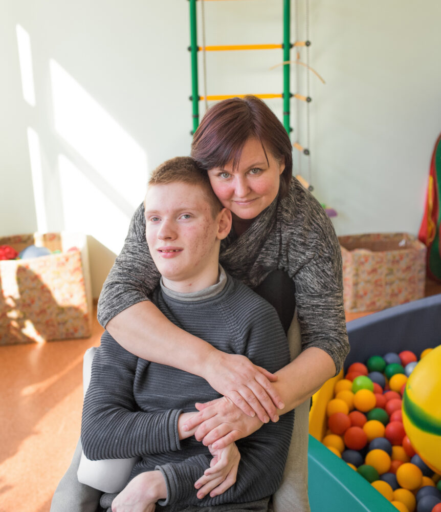 White red-haired teenage boy in a wheelchair with his caretaker lovingly wrapping her arms around him from behind - they are in a physical therapy space with a ball pit, climbing equipment