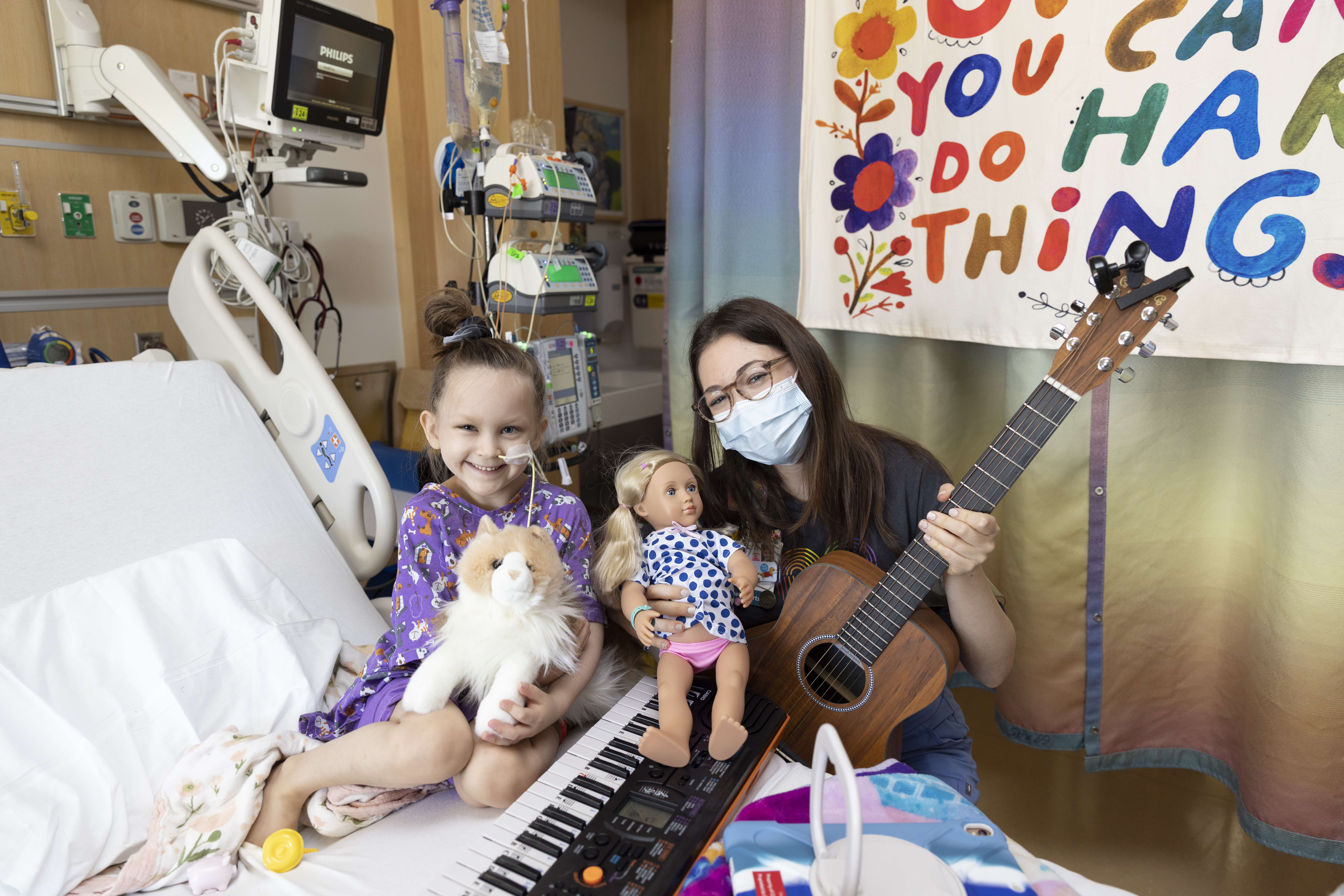 A young patient poses with her musical therapist in her hospital room. There is a keyboard on the bed and the therapist is holding a guitar.