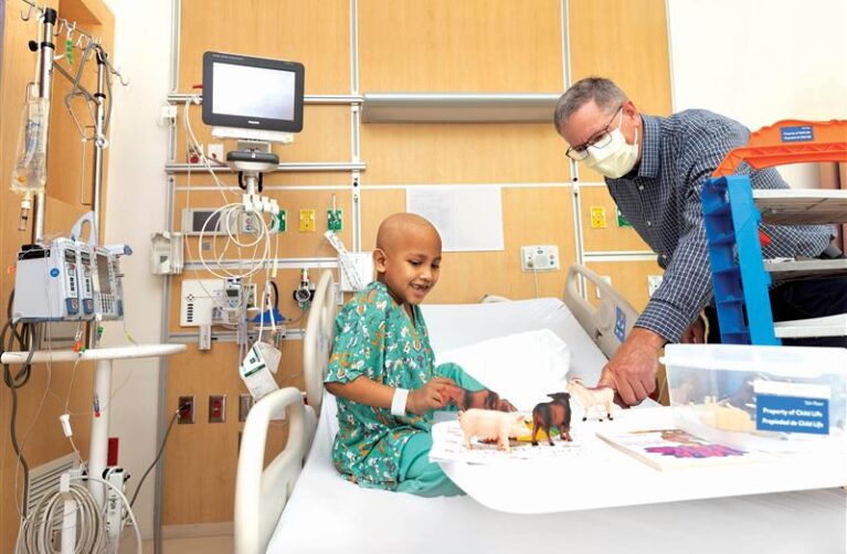Photo of a patient in his hospital bed wearing scrubs and a chaplain playing with him.