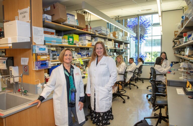 Two female doctors stand side by side in a lab. Each is wearing a white lab coat.