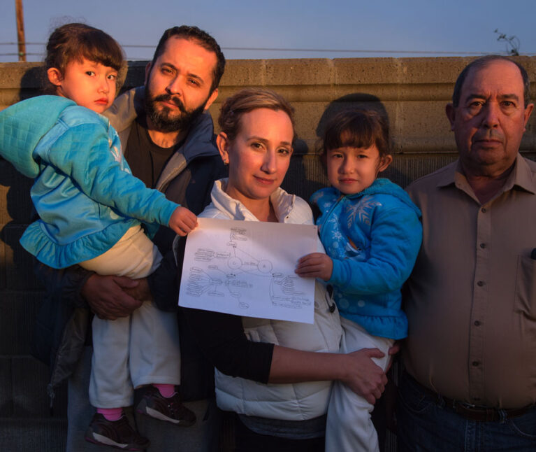 Twin girls with special health care needs are held by their parents while standing next to their grandfather. They are outside, next to a fence, their faces reflecting the sunset. The girls are holding a copy of their care map.