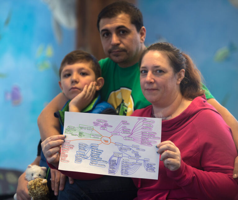 A boy with special health care needs sits on his father's lap, while his mother, seated next to him, holds up his care map.