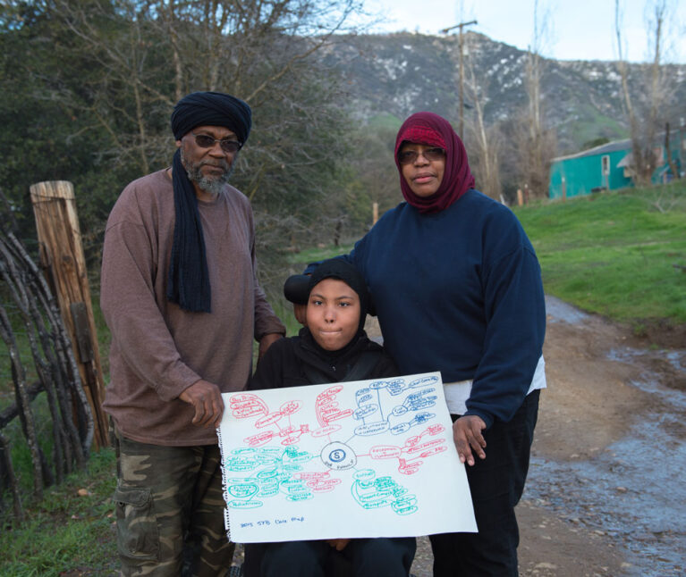 A girl with special health care needs wearing a hijab is seated in her wheelchair outside her home in a rural California town. She and her mother and stepfather are holding her care map.