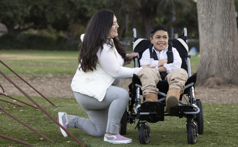 Mother kneeling to smile at her CYSHCN son who is strapped in a wheelchair