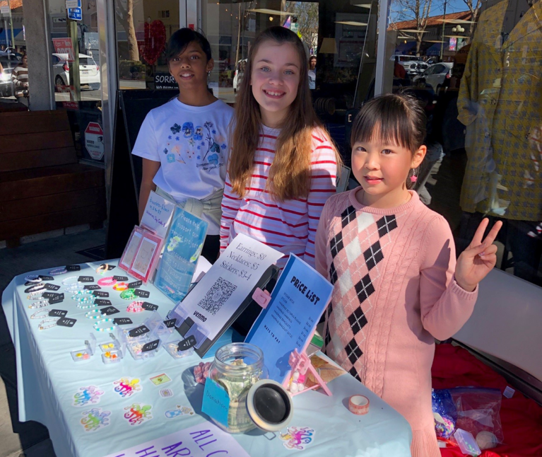 Three girls pose by their table where they are selling crafts to raise money for the children's hospital.