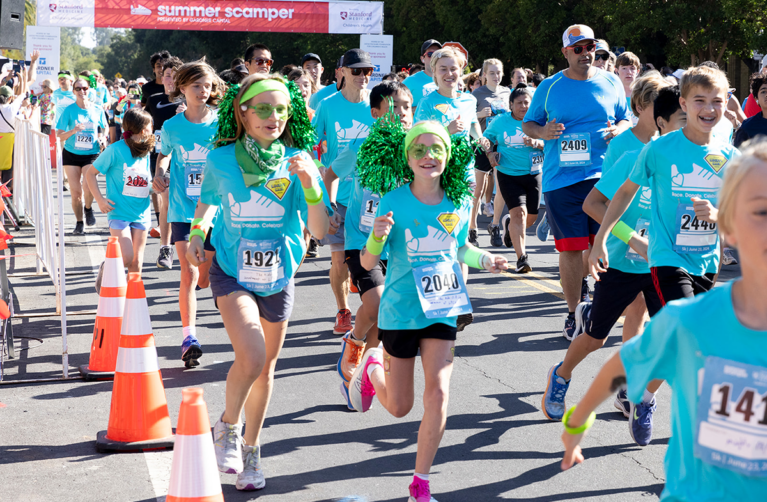 Two girls smiling at the camera among other participants as they run in Summer Scamper at Stanford University.