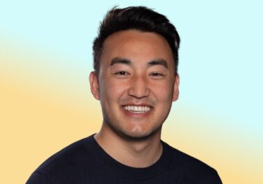 Smiling person in a dark sweater facing the camera in a studio-style headshot against a soft blue and yellow gradient background.