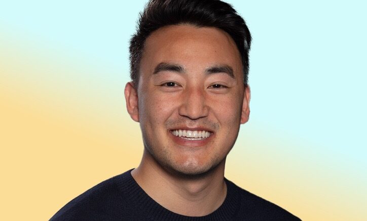 Smiling person in a dark sweater facing the camera in a studio-style headshot against a soft blue and yellow gradient background.