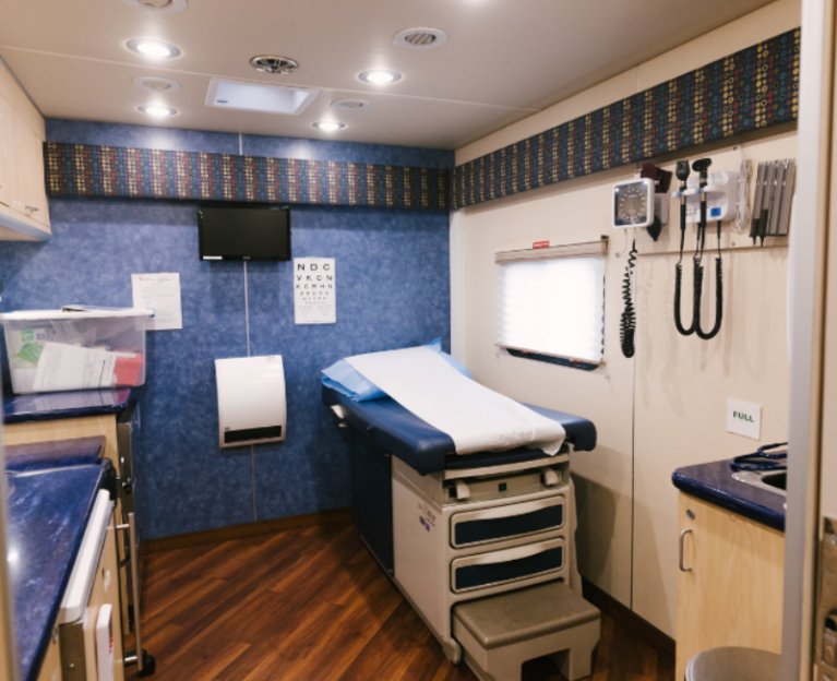 Interior of a mobile health clinic exam room with a padded exam table, wall-mounted diagnostic tools, cabinets, and medical equipment.