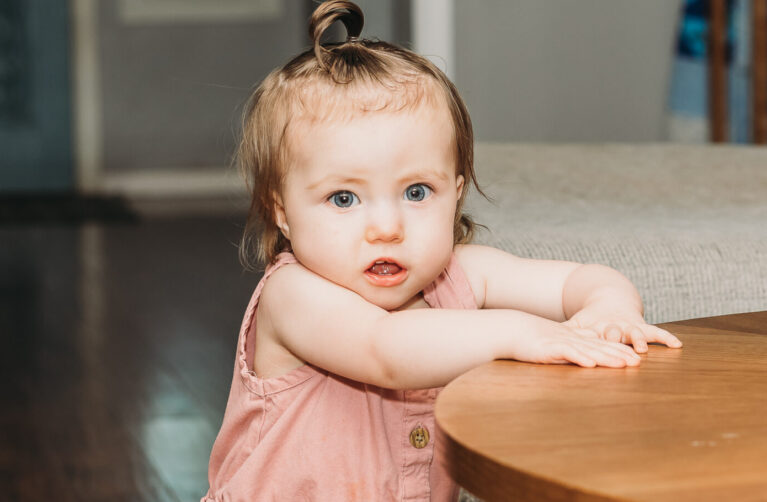 1-year-old girl, Hazel, stands by a table.