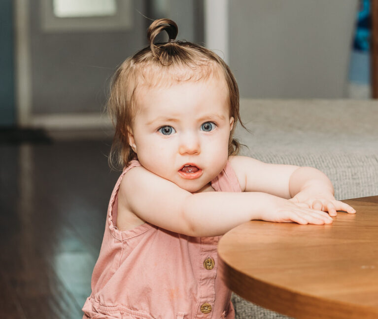 1-year-old girl, Hazel, stands by a table.