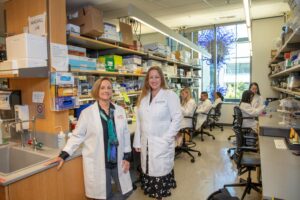 Two female doctors stand side by side in a lab. Each is wearing a white lab coat.