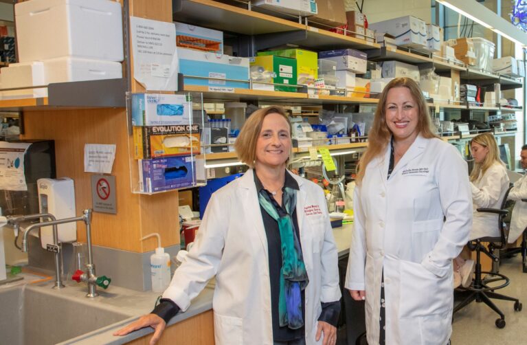 Two female doctors stand side by side in a lab. Each is wearing a white lab coat.