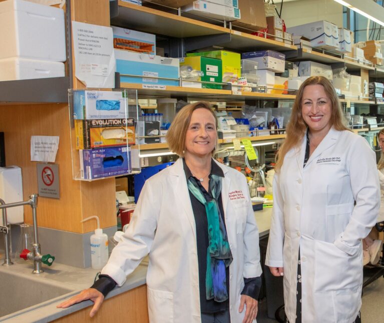 Two female doctors stand side by side in a lab. Each is wearing a white lab coat.