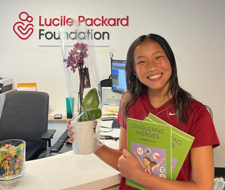 A smiling former Packard Children's allergy patient standing in front of the Lucile Packard Foundation for Children's Health logo at the Foundation's office.