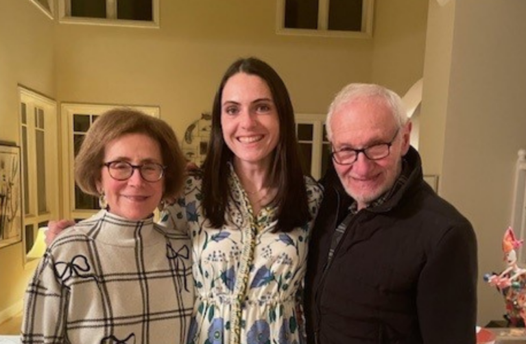 Three adults stand side by side indoors, smiling at the camera; a former packard patient in a patterned dress stands between their grandparents.