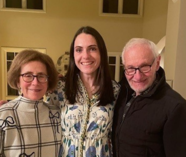 Three adults stand side by side indoors, smiling at the camera; a former packard patient in a patterned dress stands between their grandparents.