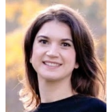 A person with straight dark brown hair parted in the center smiles at the camera. They wear a black top and stand in front of a softly blurred outdoor background.