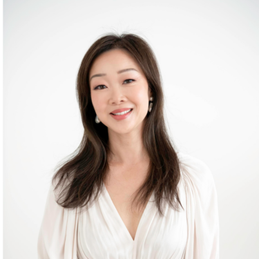 A person with long dark hair smiles at the camera against a light gray background. They wear a light-colored, long-sleeved dress with subtle pleating.