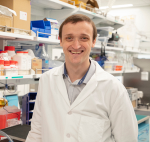 Researcher in a white lab coat standing in a laboratory workspace with lab equipment and storage shelves in the background.