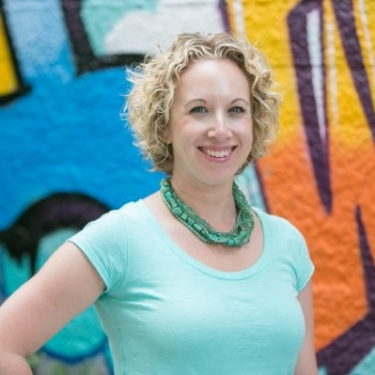A person with short curly blond hair wearing a light teal shirt and green beaded necklace stands smiling with one hand on their hip in front of a brightly colored graffiti mural.