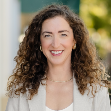 A person with shoulder-length curly brown hair smiles at the camera outdoors. They wear a light blazer, white top, gold hoop earrings, and a thin gold necklace.