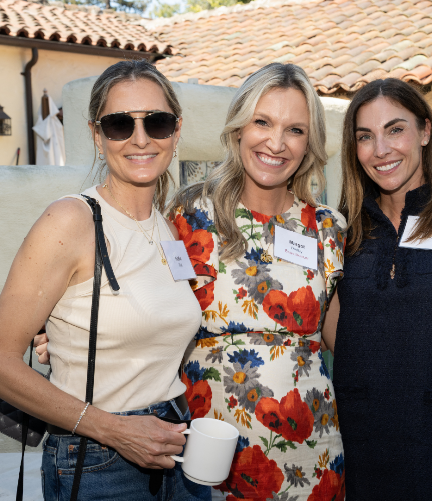 Three people stand together outdoors in front of a tiled-roof building, smiling at the camera. The person in the center wears a floral dress, while the person on the left wears a sleeveless beige top with jeans and the person on the right wears a navy dress.
