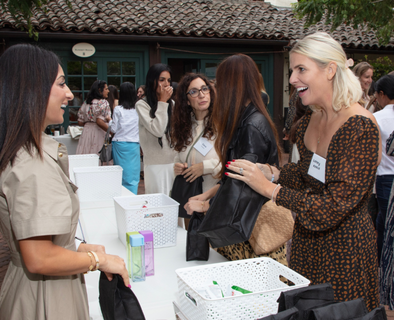 A group of people stand around a table talking and smiling at an outdoor event. Two people in the foreground face each other across the table while holding black tote bags, with other attendees conversing in the background outside a building with a tiled roof.
