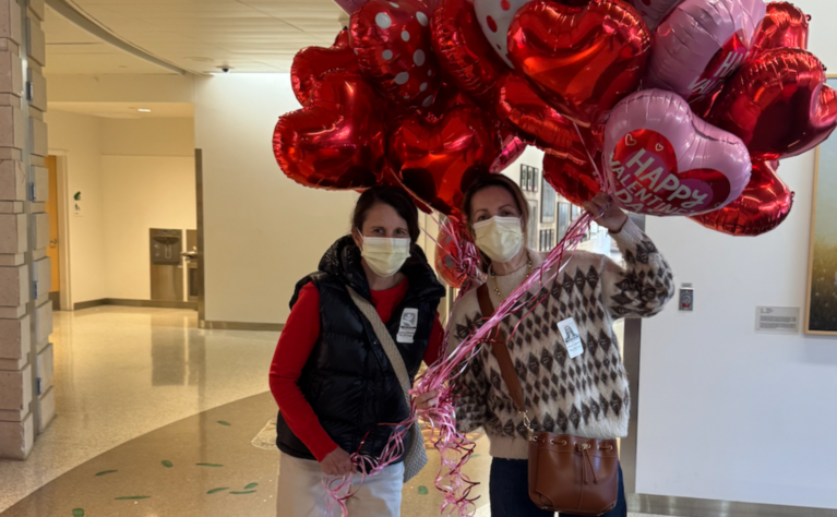Two people wearing face masks stand in a hospital hallway holding a large cluster of red and pink heart-shaped balloons with Valentine messages. A piano and framed artwork are visible in the background.