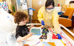 Pediatric patient engaged in art therapy with their art therapist, painting in a hospital bed and smiling in a bright, warm hospital room..