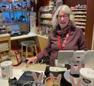 A smiling woman with gray hair and glasses stands behind the register in a gift shop, wearing a purple top and red lanyard. Shelves of cards, candy, and small gifts line the walls behind her, while an Easter-themed sign with bunny ears sits on the counter in the foreground.