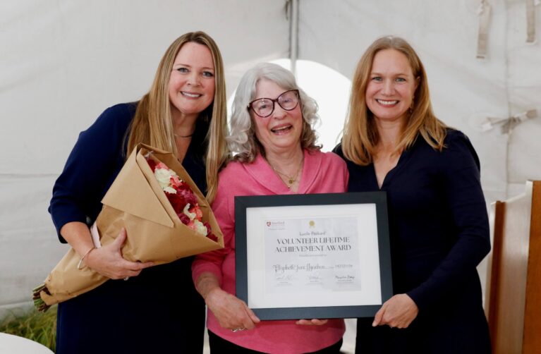 Three women smile while posing together at an award presentation. The woman in the center holds a framed Volunteer Lifetime Achievement Award certificate, while the woman on the left holds a bouquet of flowers. They stand inside a white event tent.