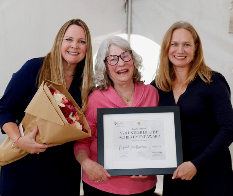 Three women smile while posing together at an award presentation. The woman in the center holds a framed Volunteer Lifetime Achievement Award certificate, while the woman on the left holds a bouquet of flowers. They stand inside a white event tent.