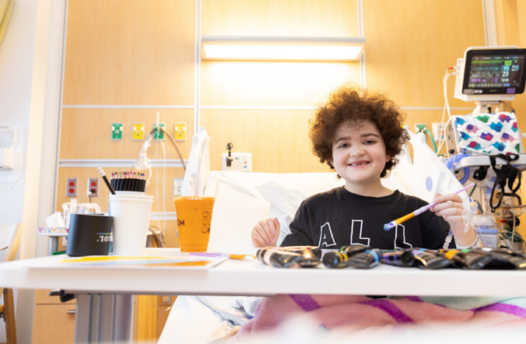 Pediatric patient engaged in art therapy, painting in a hospital bed and smiling in a bright, warm hospital room..