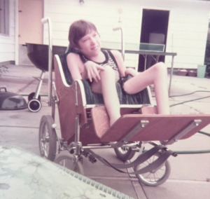 A child sits outdoors in a custom-built wheelchair, wearing a tank top and shorts. 