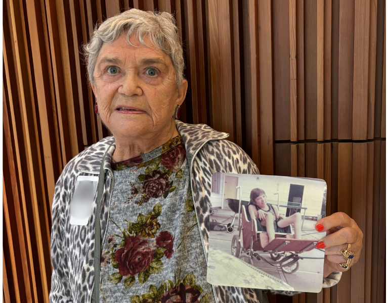 An older adult smiles while holding up a printed photograph of their late child in their right hand, presenting it toward the camera. The focus is on the photograph and the person’s expression, capturing a moment of remembrance and connection.