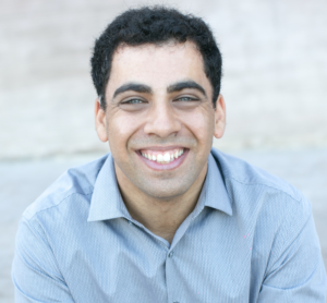 Headshot of a smiling man in a light blue button-down shirt against a soft gray background.