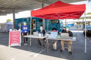 Three masked healthcare workers staff a Stanford Teen Van COVID-19 testing site under a red canopy tent in a parking lot, with the blue Teen Van visible in the background and a step-by-step testing instructions sign in the foreground.