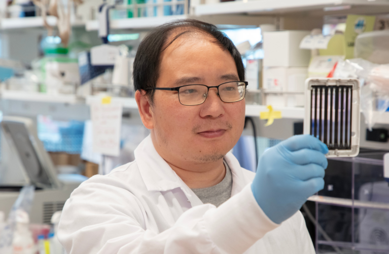 Stanford Scientist in a white lab coat and blue lab gloves observing a small piece of scientific lab equipment.