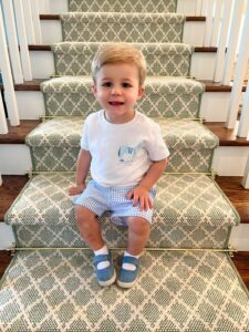 A smiling toddler boy sits on a staircase with a green patterned carpet runner.