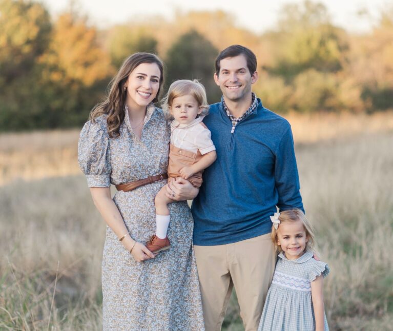A family of four poses together in an open field during golden hour, with autumn trees in the background.