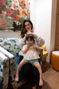 A woman with long dark hair sits in a patterned chair and fixes the hair of a small toddler girl sitting on her lap, who holds a blue toy. A colorful painting hangs on the wall behind them.