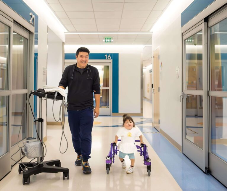 A smiling clinician in scrubs walks down a bright hospital corridor alongside a small toddler girl using a purple pediatric walker.