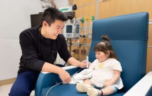 A smiling clinician in scrubs leans toward a small toddler girl seated in a large blue medical chair, gently attending to her arm as she looks on.
