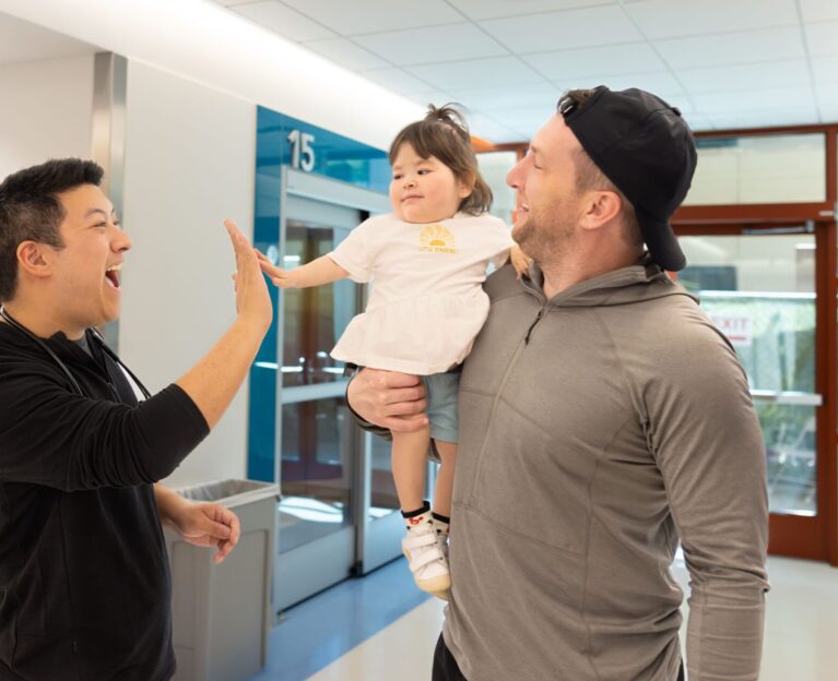 A clinician shares a high five with Audrey, a smiling toddler being held by her father, in a bright hospital hallway at Lucile Packard Children's Hospital Stanford.