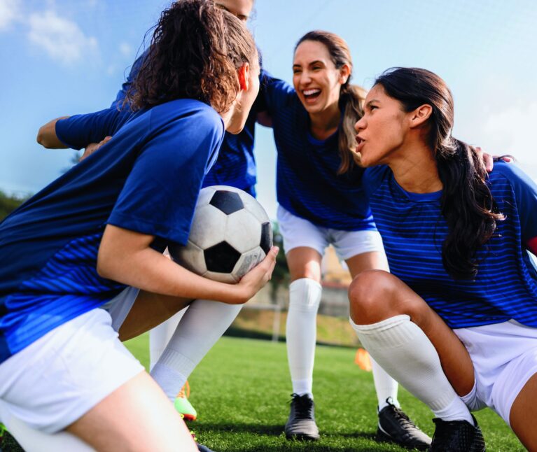 Three female soccer players in blue uniforms huddle together on a grass field, laughing and cheering, with one player holding a soccer ball.