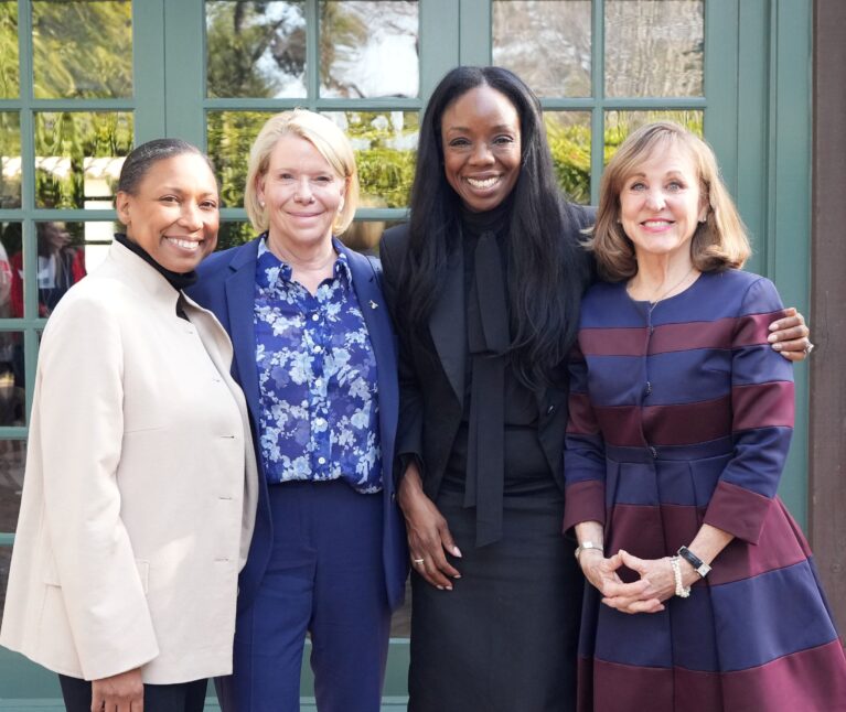 Four women smile together for a photo outdoors in front of green-framed windows. From left to right: a woman in a cream blazer, a woman in a blue suit with a floral blouse, a tall woman in an all-black outfit with a bow-tie blouse, and a woman in a navy and burgundy striped jacket. The group poses closely together in a warm, celebratory manner