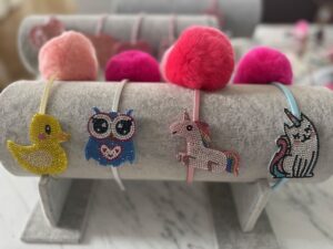 Four rhinestone-embellished headbands displayed on a gray velvet jewelry stand, each topped with a fluffy pom-pom. The headbands feature sparkly animal charms: a yellow rubber duck, a blue owl with a heart, a pink unicorn with a rainbow mane, and a silver unicorn. Pom-poms range from light pink to hot pink and coral.